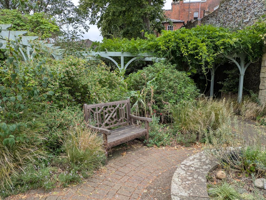 A cozy urban garden scene featuring a weathered wooden bench positioned on a curved brick pathway. Surrounding the seating area are lush, dense shrubs and ornamental grasses with feathery, beige-tinted plumes. In the background, a light blue painted metal arbor with decorative curved supports supports a canopy of vibrant green foliage, including climbing plants and vines. To the right, there is a textured stone wall, and beyond the garden, the upper floors of red brick buildings are visible under an overcast sky. The garden appears well-maintained with a mix of textured plants, trimmed hedges, and gravel areas, demonstrating thoughtful landscaping work by Gardeners Stockwell, aligned with the courtyard revamp case at Larkhall Park.