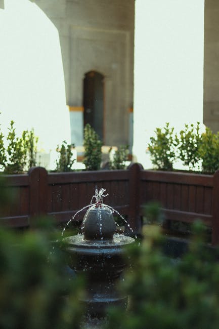 A close-up view of a small garden fountain featuring water gently jetsting from the top into a basin below, surrounded by lush green plants with small, rounded leaves. In the background, there is a wooden fence with evenly spaced, trimmed hedges behind it, along with a light-colored building with a white archway and a door, all bathed in natural daylight. The garden area appears well-maintained, showcasing flowering shrubs and neatly edged flower beds, with a smooth lawn or soil surface visible. The scene captures the tranquil atmosphere typical of a landscaped courtyard, reflecting gardening and outdoor maintenance activities carried out by Gardeners Stockwell as part of their landscaping work for the courtyard revamp case by Stockwell gardeners Larkhall Park.